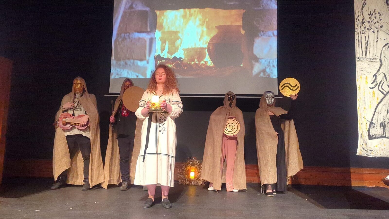 The Trypillian Tarot ensemble in hooded burlap cloaks and gold masks on the La Mama Courthouse stage, gathered around a lit lantern beneath a projection of Trypillian pottery.