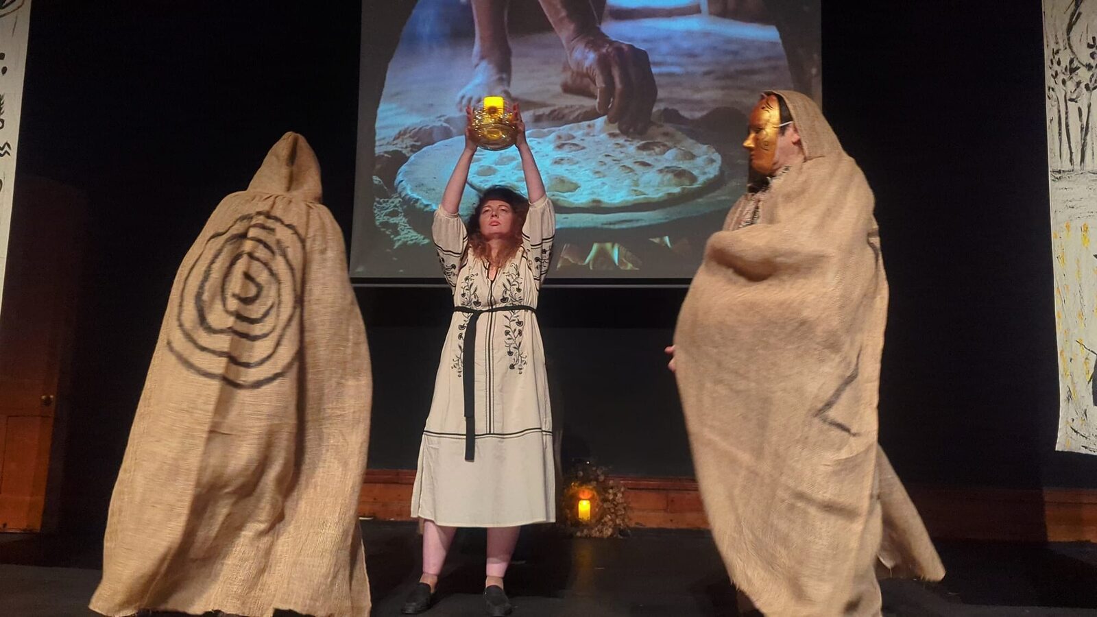 A performer in a white embroidered dress raises a glowing vessel overhead, flanked by two cloaked figures painted with charcoal spirals.
