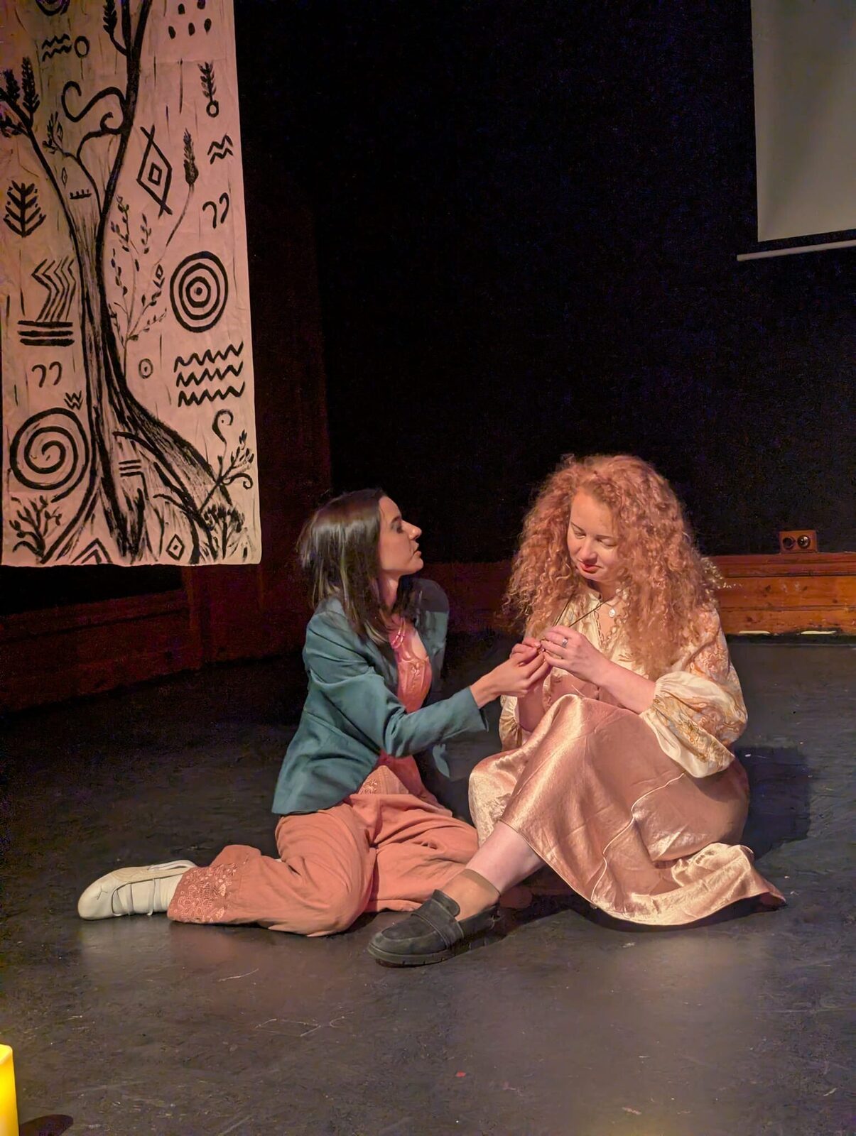 Two performers sit close together on the stage floor, hands joined, in a quiet moment between scenes.