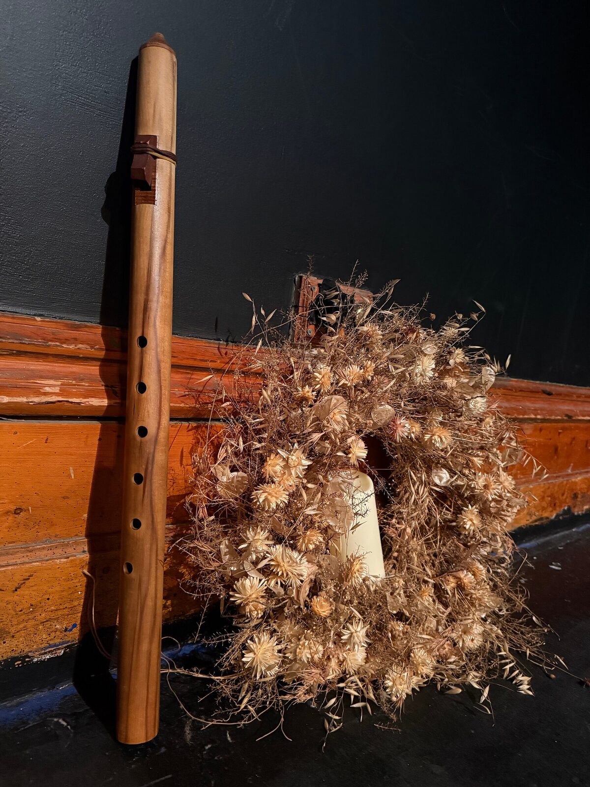 A wooden flute resting against a dried wheat and thistle wreath, lit warmly against the dark stage wall.