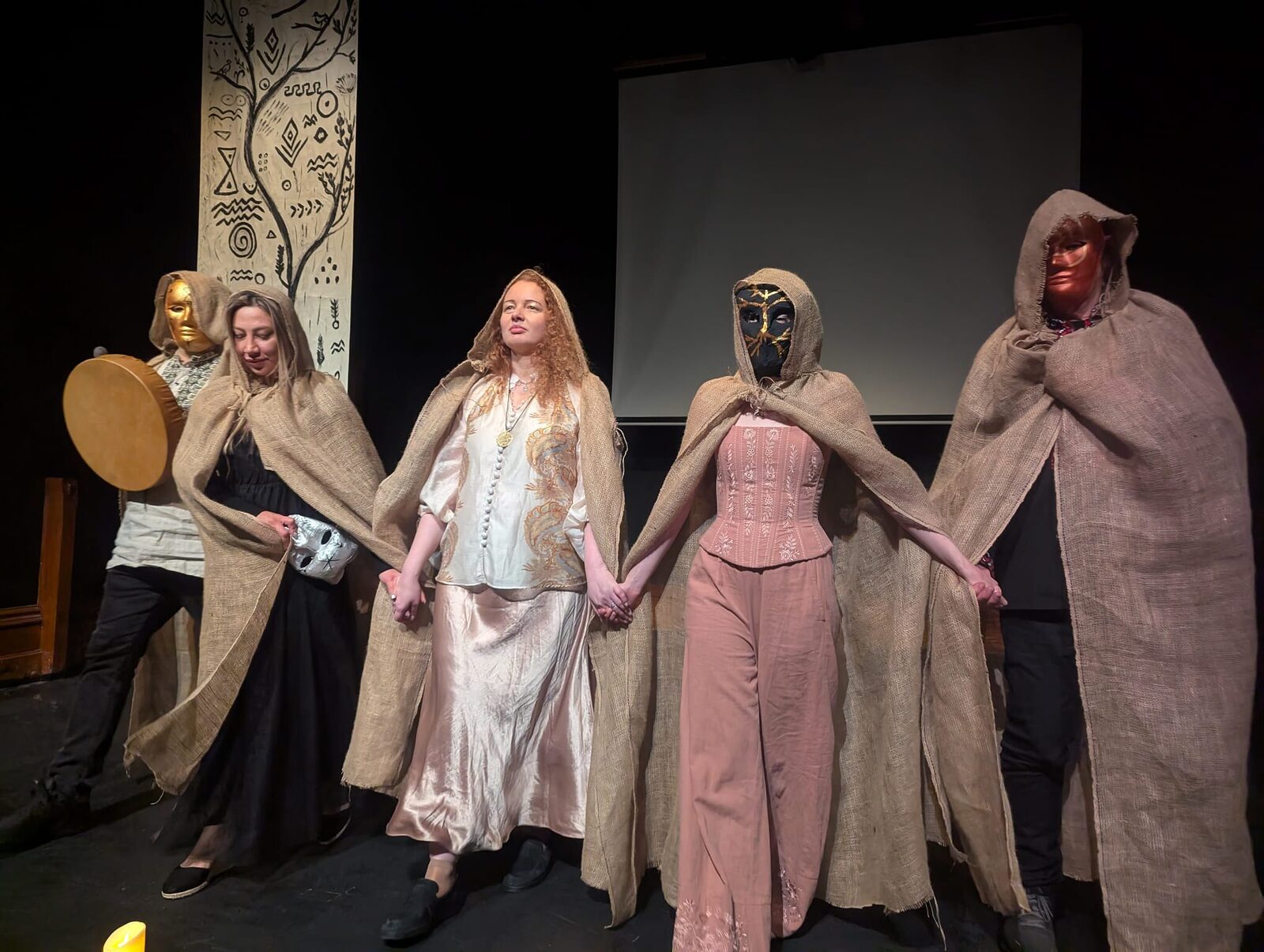 The full cast stand hand in hand across the stage in costume, cloaks and masks lowered.